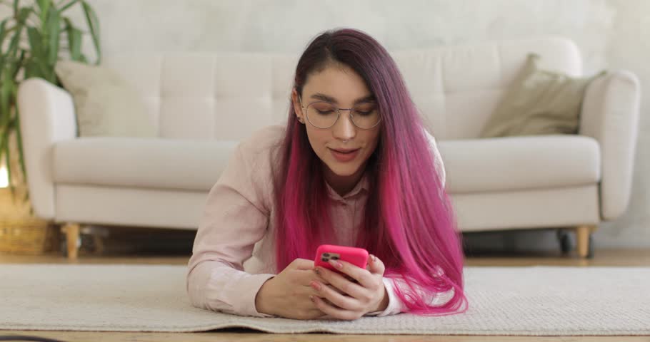 A young caucasian woman with colored hair is lying on a carpet in a white living room and using a smartphone. Chatting, internet communication. Modern lifestyle. | Shutterstock HD Video #1100454157