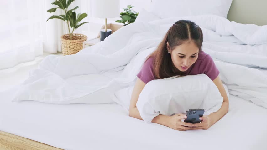 Happy Asian woman laying in bed using a smart phone with a finger touching the screen in the morning. Women use device for social networks, chat or online shopping from smart phones. | Shutterstock HD Video #1100458147