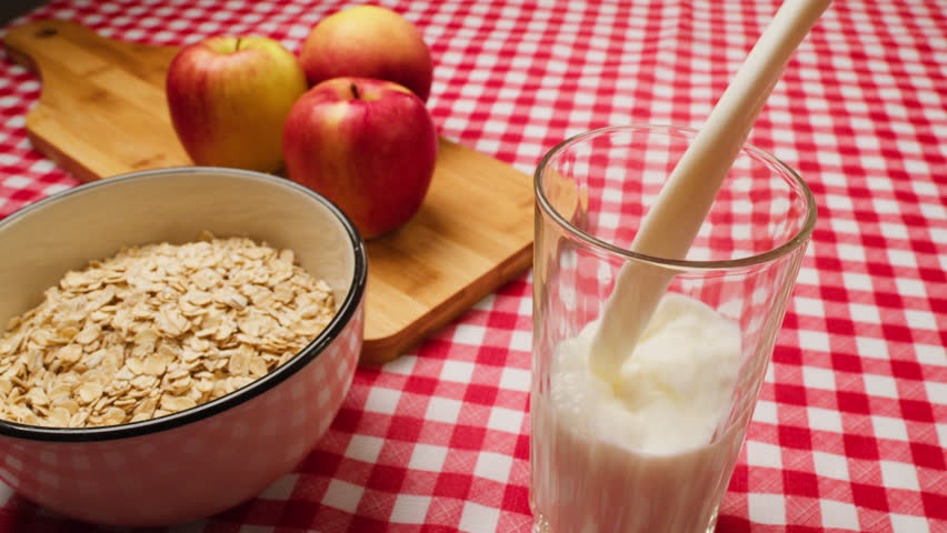 Milk pours out into a glass. Glass with yogurt, dairy products concept. Pouring milk into a glass on a table with a red tablecloth. Healthy breakfast in the morning. Cereals and milk | Shutterstock HD Video #1100458319