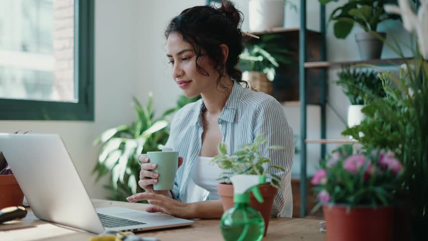 Video of smiling woman drinking a cup of coffee while working with her laptop in greenhouse. | Shutterstock HD Video #1100460553
