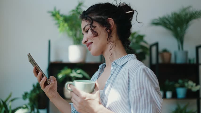 Video of smiling woman drinking a cup of coffee while using her mobile phone in greenhouse. | Shutterstock HD Video #1100460567