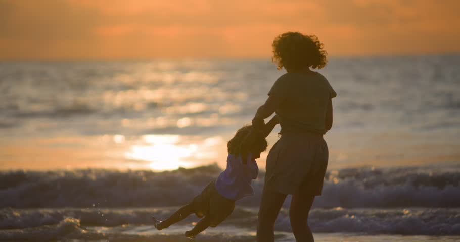 Mother spins her son, holding his hands while stand on shore by sea and waves. Warm and soul-warming memories of childhood that will remain in memory of little boy. Mommy plays with her baby at sunset Royalty-Free Stock Footage #1100460743
