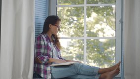 Young woman reading book near window. Beautiful female read book sitting on windowsill at home. Female student study at home. Girl with novel book relaxing on window sill. Realtime - Powered by Shutterstock - Get 15% off with code: PIKWIZARD15