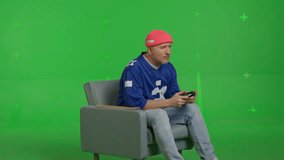 Close-up of a white man dressed in a blue sports jersey and beanie sitting in a chair and playing with a joystick in a video game on TV on a green screen background, chroma key, side view