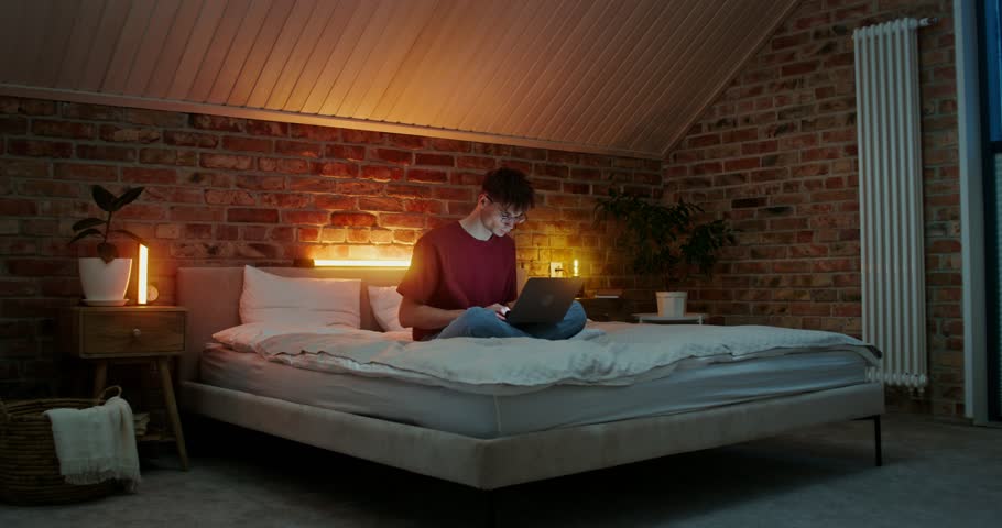 A young man with glasses uses a laptop while sitting on a bed in a lotus position. Video filming in the home interior