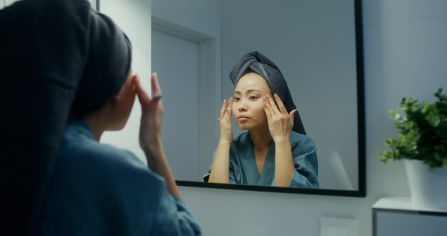 A young Asian woman in a bathrobe, with a towel on her head, makes a facial massage with her hands, looking in the mirror in the bathroom