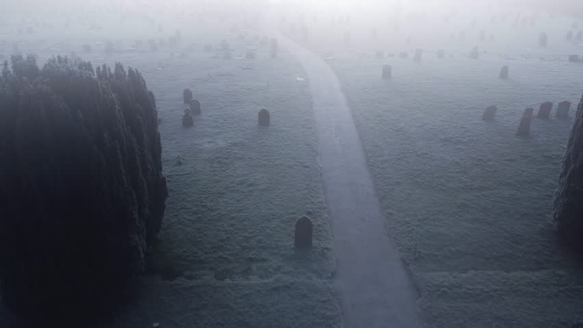Aerial drone shot over a foggy frozen Thetford cemetery on London Road showing snow filled grave yard on both sides of a path.