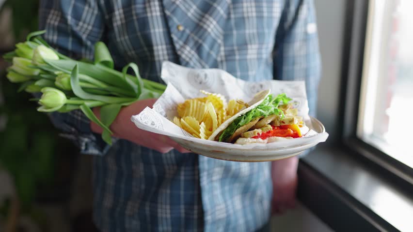 Chef holding a freshly made Italian sandwich on a plate in an Italian deli restaurant. Italian deli sandwich on display with flowers and a napkin beside it.