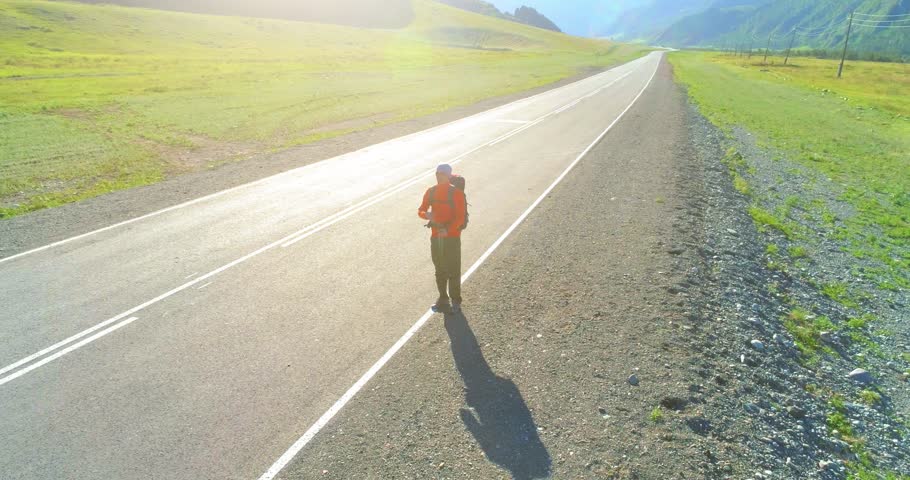 Aerial 4K UHD view. Low altitude flight over hitchhiker tourist walking on asphalt road. Huge rural valley and sunny meadow at summer day. Backpack hiking guy.