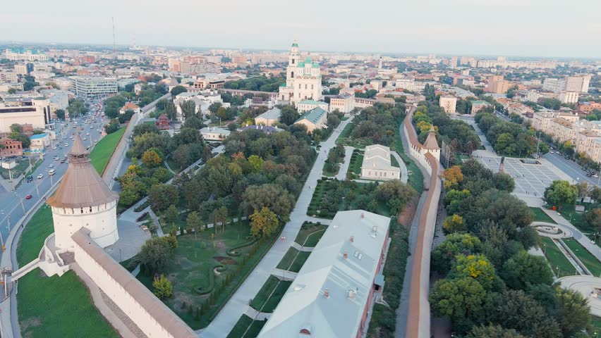 Astrakhan, Russia. The Astrakhan Kremlin dominates the center of the frame, its massive white-stone fortress walls and towers enclosing a complex of historic structures crowned by the five-domed Cathedral of the Assumption with its distinctive green cupolas. The camera pulls back smoothly from an elevated aerial position, gradually revealing the full triangular perimeter of the 16th-century citadel and its integration into the surrounding urban fabric. The fortress sits on elevated ground with formal gardens and pathways visible inside the walls, while modern city streets and residential blocks extend outward in all directions. The scene is captured during the golden hour of late afternoon, with warm, soft light casting long shadows across the landscape and highlighting the contrast between the pale stone of the Kremlin and the dense green canopy of the trees within and around the complex. The overall atmosphere is serene and majestic, emphasizing the enduring presence of this historic landmark within a contemporary cityscape. The stone walls, built using recycled Tatar brick from the ruins of Golden Horde cities, still retain their original defensive character with crenellated parapets and angular towers. Departure of the camera