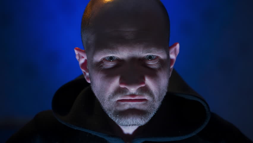 closeup portrait of a stern-looking man with under face light, blue background, exudes an unfriendly, serious vibe. dramatic visual scene. criminal man is defiantly staring into the camera. scary gaze Royalty-Free Stock Footage #1100482739