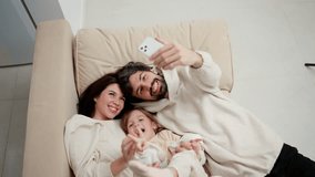 Young family are making a selfie, posing while laying on a sofa in a living room, high angle view