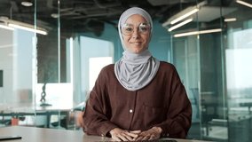Webcam view of smiling young muslim businesswoman in hijab with glasses looking at the camera and talking by online meeting conference video call having conversation at modern office workplace indoors