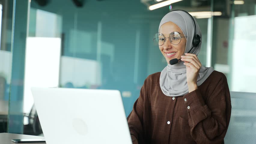 Positive young muslim businesswoman support manager in hijab with wireless headset looking at laptop screen camera and talking by online meeting conference video call at workplace in modern office Royalty-Free Stock Footage #1100486285