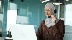 Positive young muslim businesswoman support manager in hijab with wireless headset looking at laptop screen camera and talking by online meeting conference video call at workplace in modern office