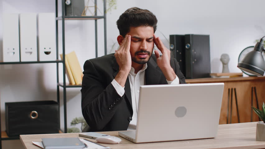 Tired ill indian businessman suffering from headache problem tension and migraine, stress at home office desk workplace. Bearded freelancer broker worker man works on laptop notebook. Overworking | Shutterstock HD Video #1100486809