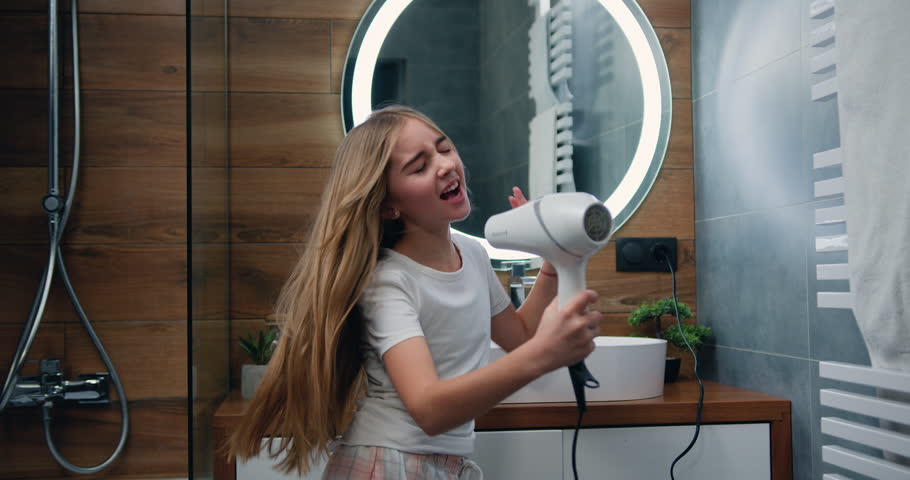 Beautiful blond little girl in bathrobe singing a song and dancing with a hair dryer. Child sings and dances in a shower after taking shower in bathroom. Royalty-Free Stock Footage #1100488609