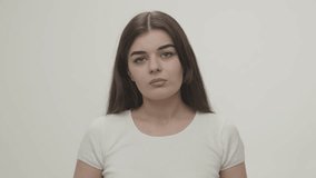 Video of a young brunette feeling sad and looking at the camera. Portrait shot of a young woman with a sad look who stands in front of a white background.