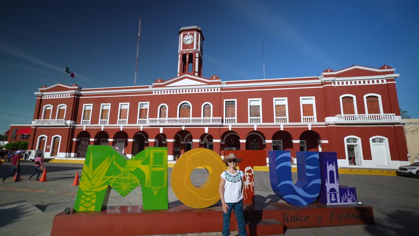 Happy smiling mature senior tourist woman wearing ethnic attire, holding out her arm as if presenting the Motul sign in front of the cities Municipal Palace.