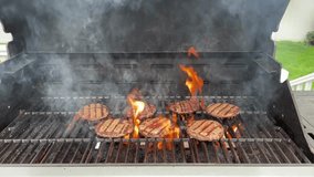 The top down, close up view of several hamburgers cooking on the grill. Someone is flipping the burgers and the flames are high.