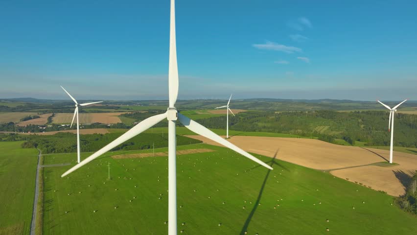 A complex of wind power plants installations in a hilly landscape. An aerial view across the variously distant wind farms in operation. Spinning propellers, Blue sky, farmed countryside. 
