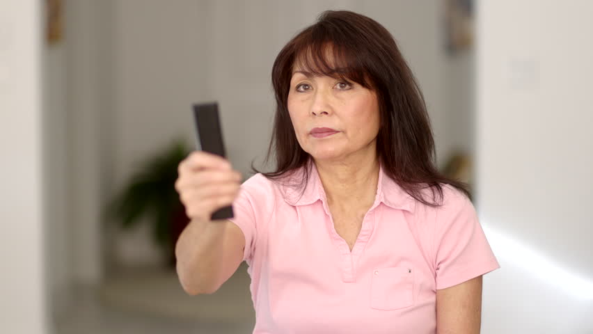 Front view beautiful stylish woman long hair in pink top changing television channels with remote control sitting in comfortable home living room. Attractive middle age asian has happy lifestyle.