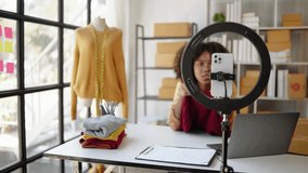 Female African designer and online seller is live streaming broadcast for selling new clothes and receiving the order from customer  and packaging product in workspace.