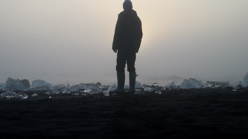 Slow motion back view shot of a man surrounded by ice formations on the shore of Diamond Beach with a backdrop of the rushing waves during sunset