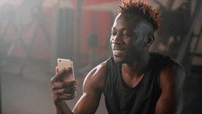 Happy African American man having a break during the training in the gym and using phone to check newsfeed in social media