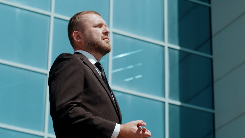Close-up outdoor portrait of confident middle aged bearded business man in suit posing outdoor with folded arms, looking aside near office building. Successful career, corporate