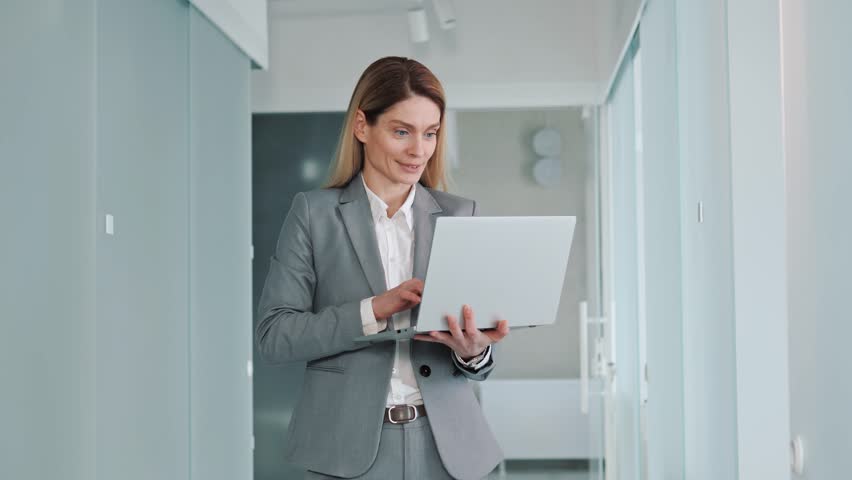 Beautiful Specialist in Formal Grey Suit Standing in Hallway Office, Holding Laptop Computer, Looking at Screen, Smiling Charmingly Typing Message. Working on Design, Data Analysis, Plan Strategy.  | Shutterstock HD Video #1100511419
