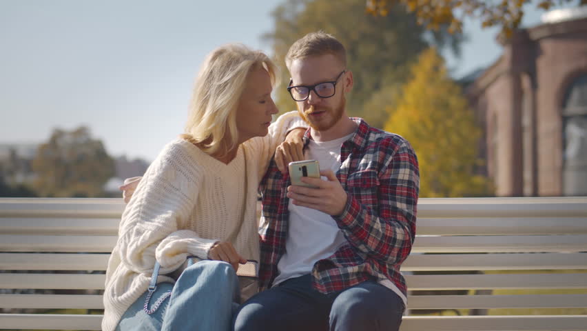 Young man show photos on smartphone to mature mother. Senior woman with adult son use cellphone outdoors. Mom and son with mobile phone spend time together in city. Realtime. 