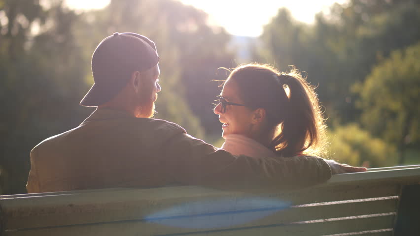 young attractive lovers sitting on bench in the park. Back view of man and woman hug outdoors. Boyfriend and girlfriend relax on bench. Couple on date. Realtime