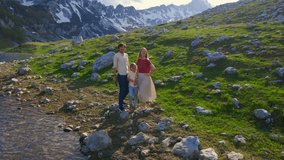 A young family captures their outdoor adventure using a drone camera. The father, mother, and their son stand in a breathtaking mountain valley with snow-capped peaks in the background, dressed