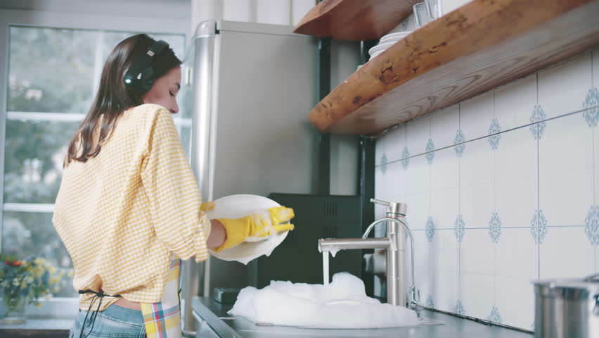 Authentic happy caucasian woman in headphones washing the dishes and dancing in a modern kitchen. Young housewife listening music, having fun while doing the washing-up. Realtime Royalty-Free Stock Footage #1100514853
