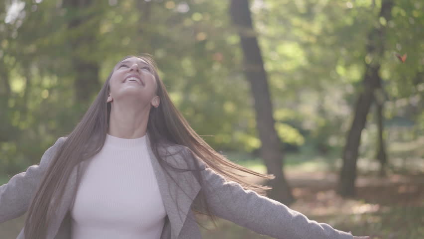 Medium shot - Slow motion of a young, brown-haired, female in a gray coat, spinning around in a forest in daylight, while autumn leaves are falling on her | Shutterstock HD Video #1100515721