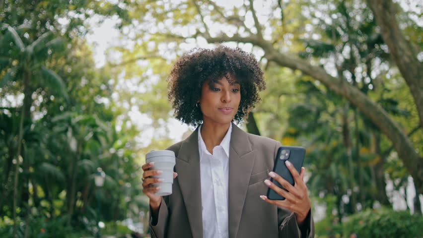 Relaxed woman talking phone video call walking city park hold paper coffee cup closeup. Attractive confident african american business lady speaking online outdoors. Pretty girl use smartphone camera Royalty-Free Stock Footage #1100516261