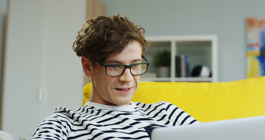 Close up of the young Caucasian man in glasses typing while chatting on the laptop computer on the couch in the living room.