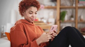 Beautiful happy young african american girl sitting on sofa in living room at home using mobile phone. Online communication distance friends online in mobile chat app using smartphone video chat.