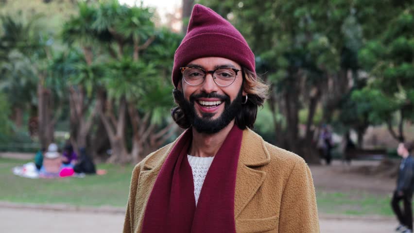 Front view of young adult man with goggles and beard, smiling and looking at camera, wearing a beanie hat and scarf in a park outdoors with happy and sucessful attitude. Portrait concept. Slow motion
