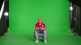 Caucasian man dressed in a red sports jersey and beanie sitting in a chair and playing with a joystick in a video game on TV on a green screen background, chroma key, side view