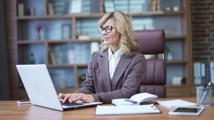Portrait of smiling mature businesswoman in suit and glasses typing on laptop at desk at workplace in office. Confident female financier or owner looking friendly at camera. Accountant at work Royalty-Free Stock Footage #1100530241