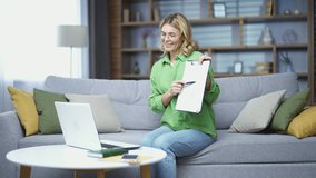 Confident mature female teacher conducting online lesson using laptop while sitting on sofa in living room. A smiling woman, speaking to students, shows a graph, a diagram she is holding in her hands
