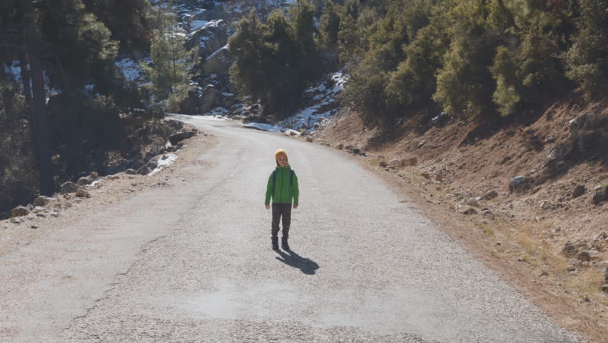 child with a backpack stands in the middle of the road in the forest and raises his hands up. hiking in the forest with children.