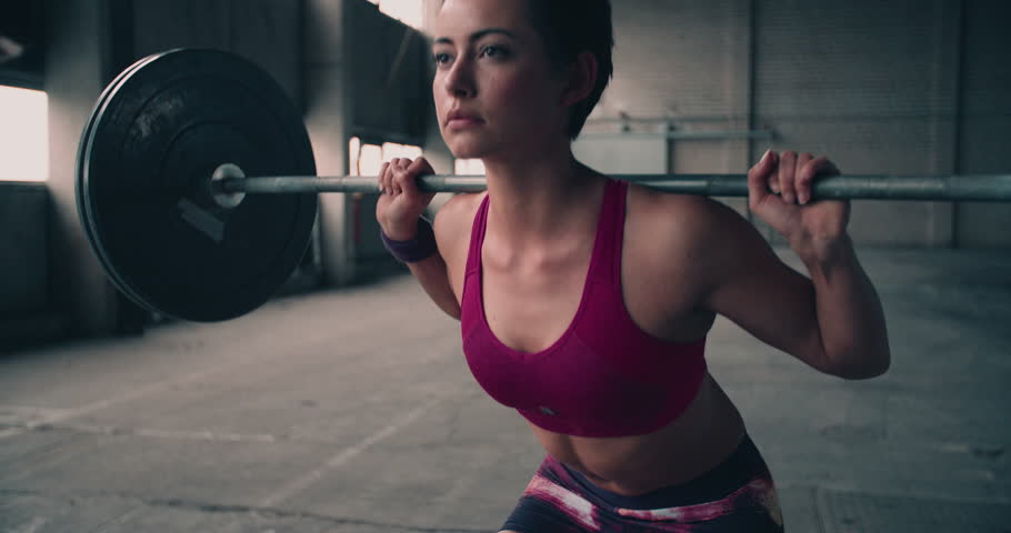 Full length image of a young woman testing her strength by holding a barbell with heavy weights on her shoulders as she squats