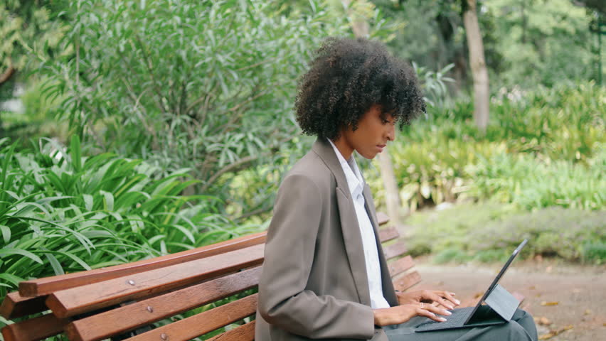 Focused woman freelancer sitting bench typing on laptop close up. Attractive african american business lady resolving work issues using computer in green park. Serious young girl doing remote job. Royalty-Free Stock Footage #1100539037