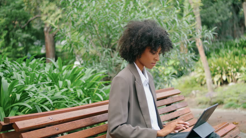 Focused woman freelancer sitting bench typing on laptop close up. Attractive african american business lady resolving work issues using computer in green park. Serious young girl doing remote job. | Shutterstock HD Video #1100539037