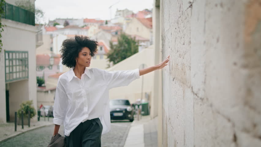 Relaxed stylish model walking alone watching city architecture wearing white shirt. Beautiful african american woman going street touching old building wall. Curly tourist enjoying cityscape outdoors. Royalty-Free Stock Footage #1100539047
