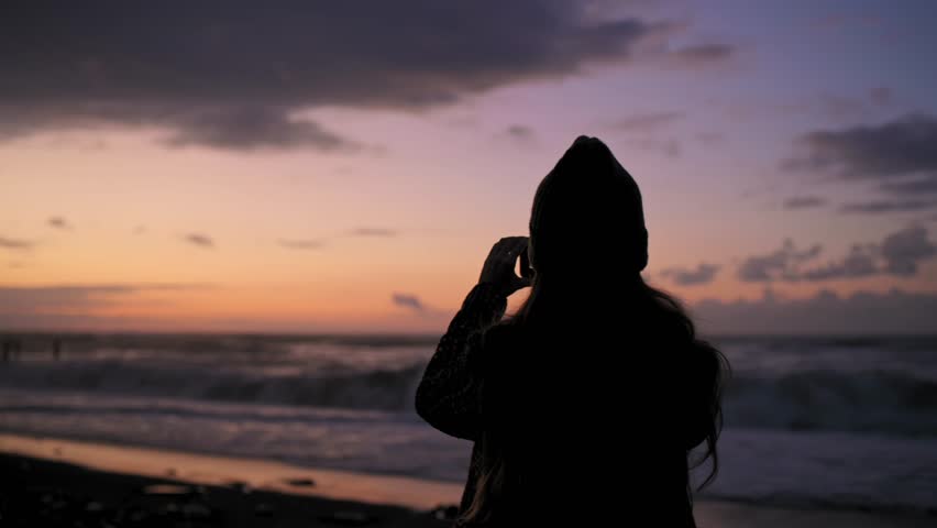 Young woman tourist takes pictures, shoots video of sunset on beach on modern smartphone. Beautiful bright sunset on the sea, ocean. Girl enjoys nature, dreams, relaxes. Hands with phone