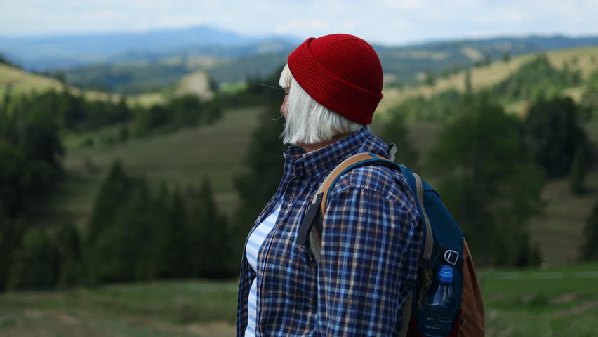 Senior woman hiker in stylish clothes, hiking backpacker traveler camper with her phone camera on the top of mountain in sunny day. Beautiful mountain landscape view. Hiking, backpacking. 4K 
